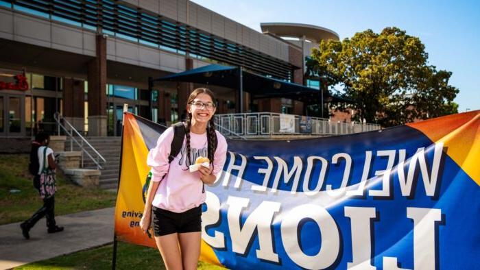 Student poses in front of a "Welcome 首页 狮子s" banner, wearing shorts and a t-shirt and holding a donut.